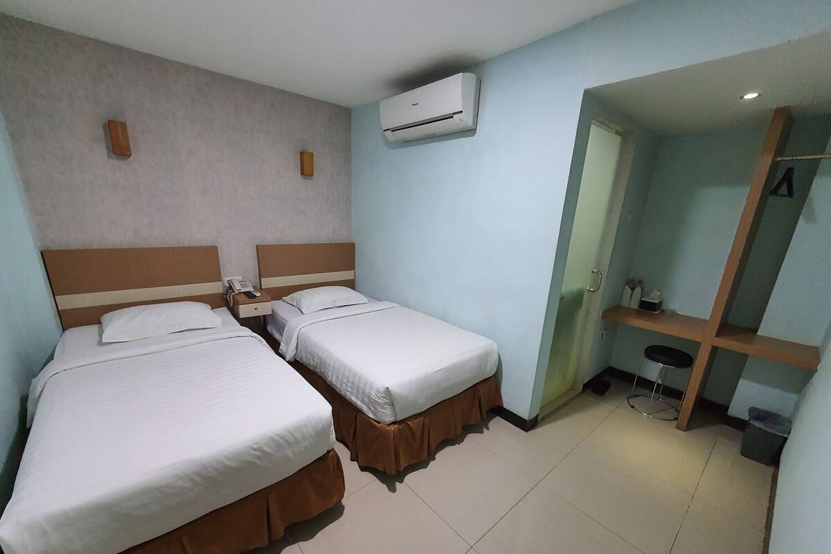 Ein Hotelzimmer mit zwei Betten, einem Schreibtisch, einem Stuhl, einer Klimaanlage und einem kleinen Badezimmerbereich.