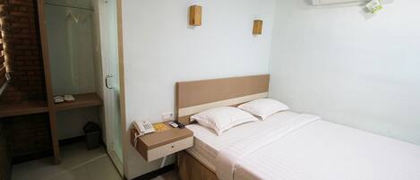 Ett hotellrum med en säng, ett sängbord med en telefon och en bok, en liten hylla, en spegel och en luftkonditioneringsanläggning.
