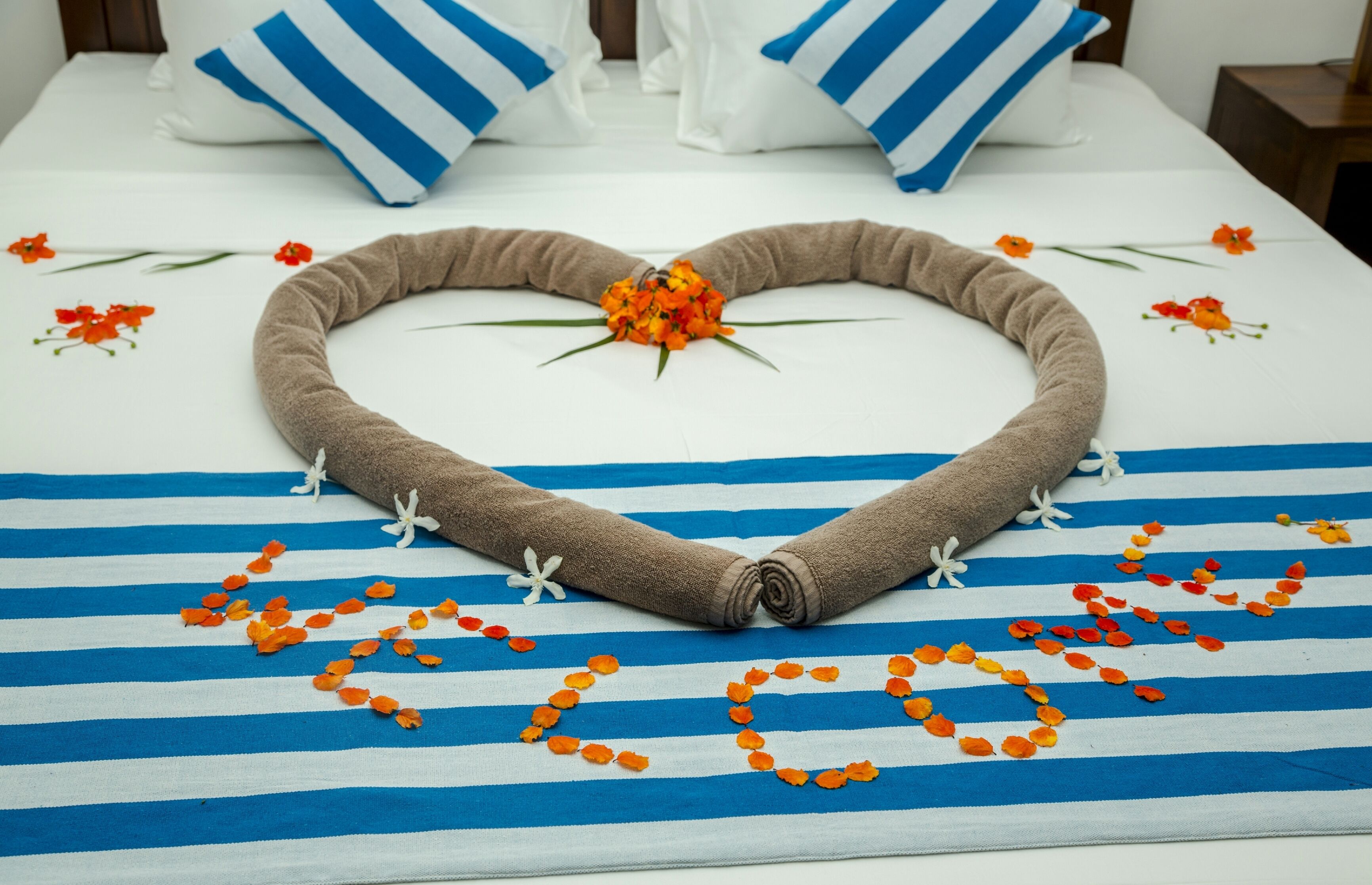 A bed with a heart-shaped towel arrangement, orange petals, and a central flower on a striped bedspread.