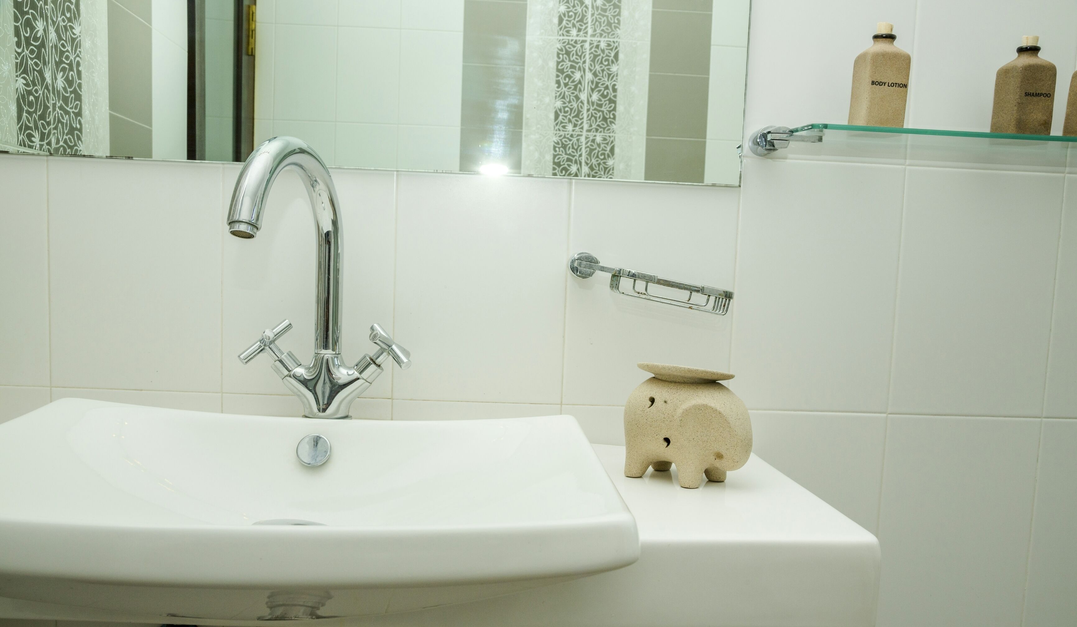 A bathroom with a white sink, chrome faucet, and a mirror above it. There is a soap dish with a soap bar on the wall, and a glass shelf with two bottles labeled "body lotion" and "shampoo".