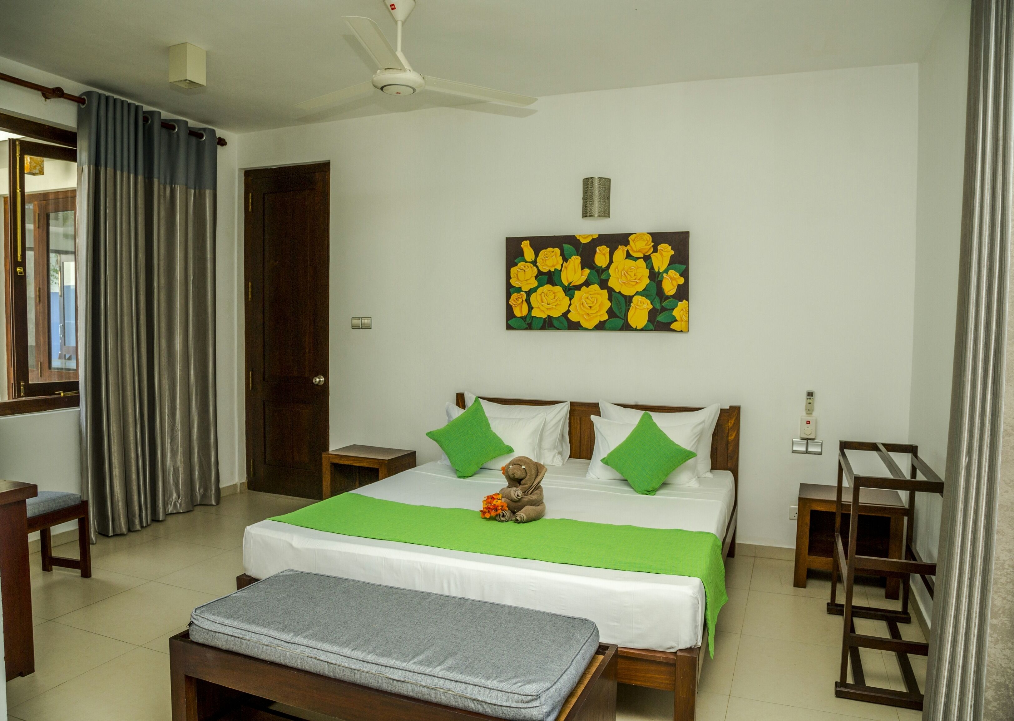 Une chambre d’hôtel avec un lit, une table de chevet, une chaise, une fenêtre avec des rideaux, un ventilateur de plafond et un tableau représentant des fleurs jaunes accroché au mur.
