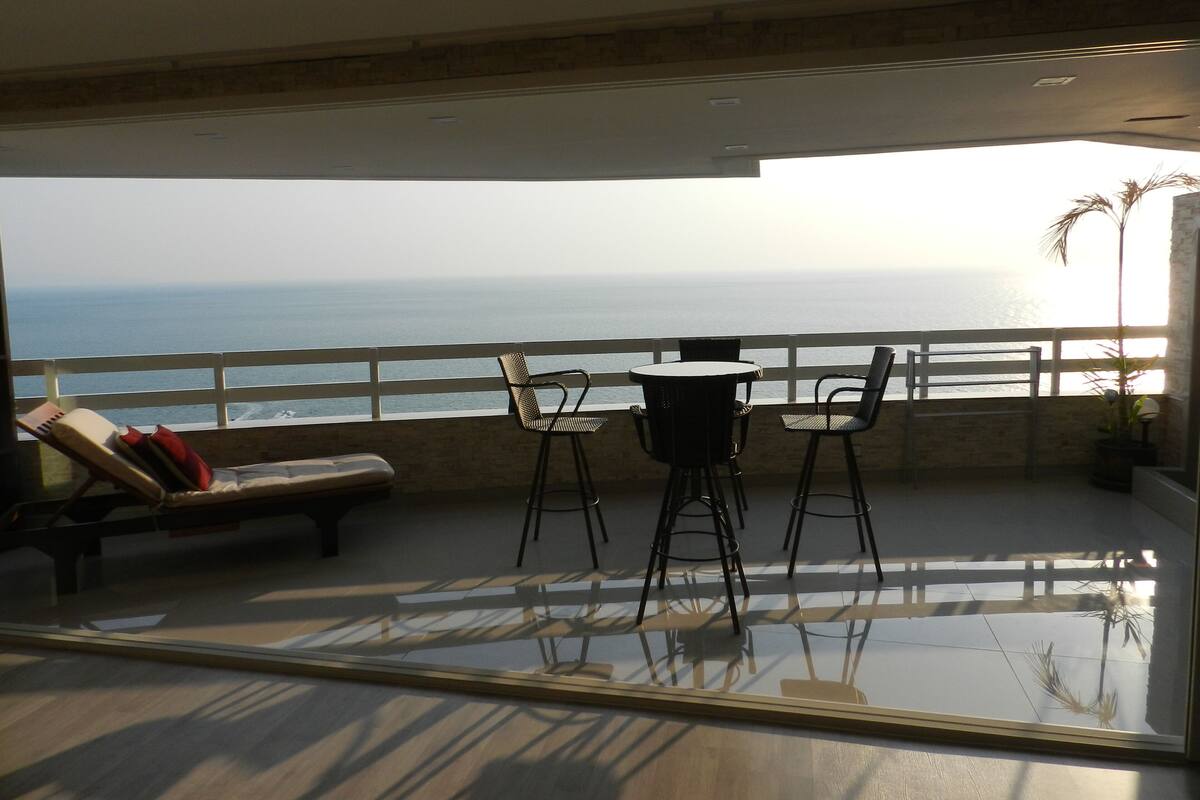 A balcony with a view of the ocean, a round table, two chairs, a bench, and a potted plant.