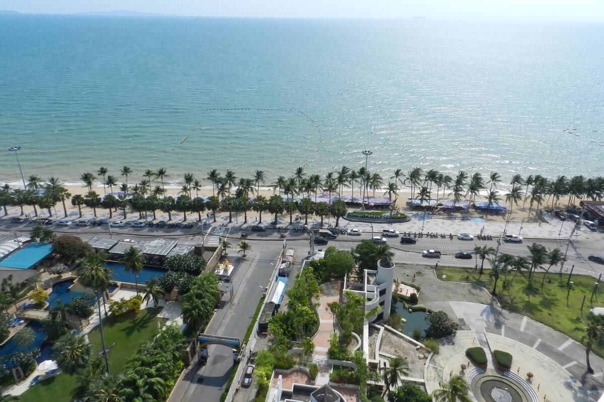 An aerial view of a coastal resort with a beach, palm trees, a swimming pool, and a parking area.