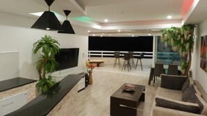 Una sala de estar moderna con una televisión de pantalla plana, un sofá, una mesa de centro y un comedor con sillas. Hay un balcón con barandilla y vistas al cielo nocturno.