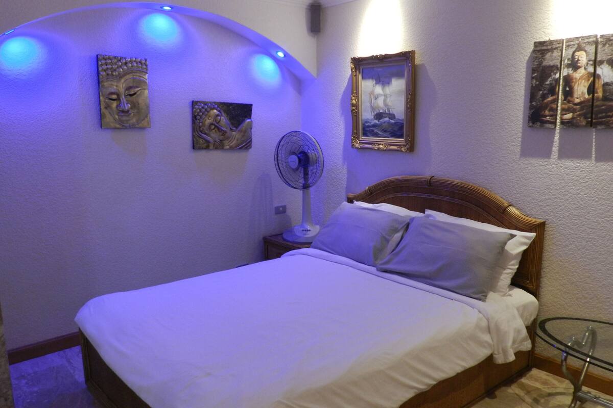 A bedroom with a bed, a bedside table, a fan, and wall art.