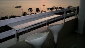 Un balcón con vista a la puesta de sol sobre el mar, con dos sillas blancas y una barandilla blanca.