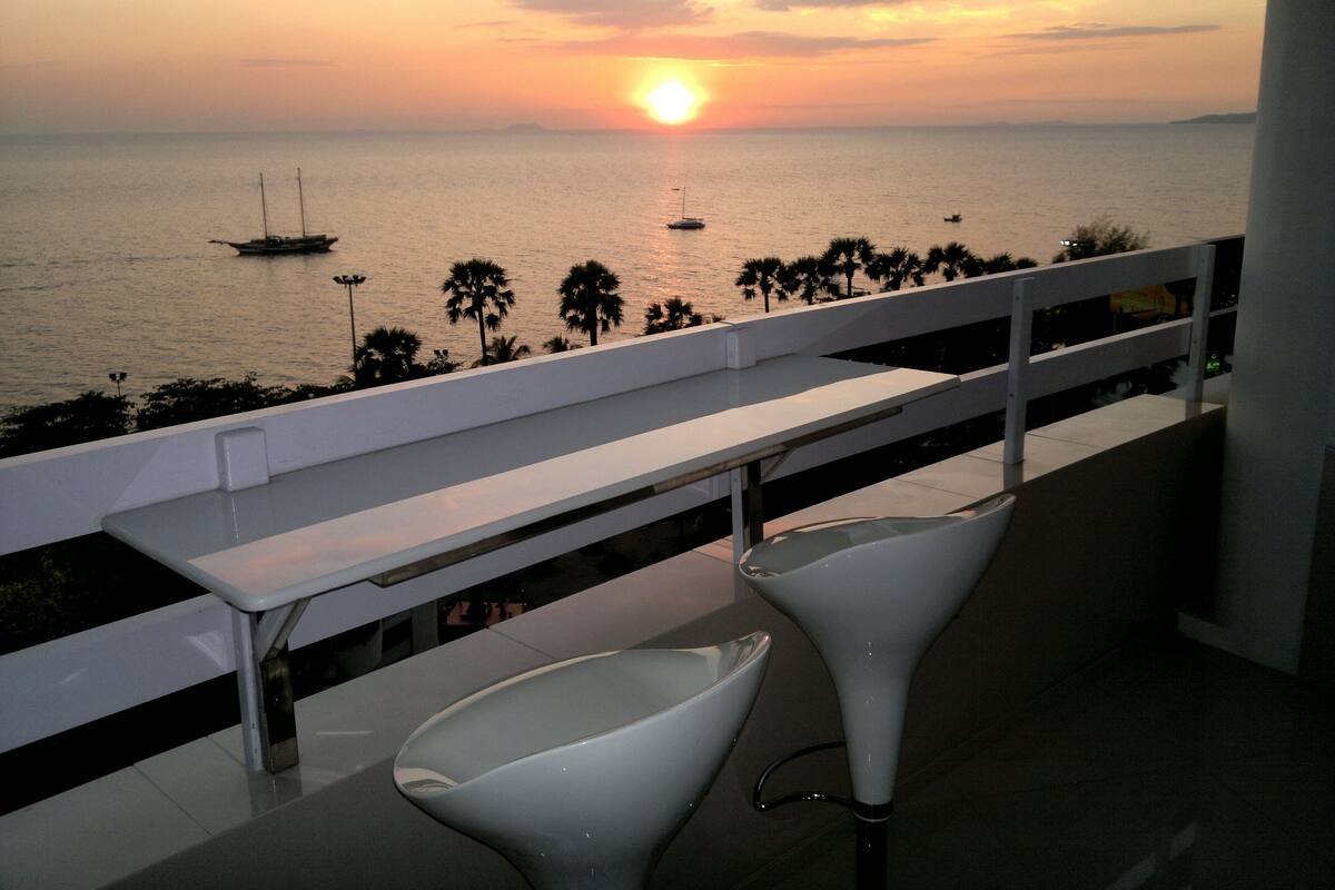 A balcony with a view of the sunset over the sea, featuring two white chairs and a white railing.