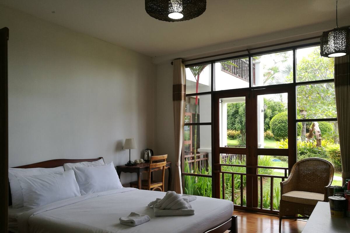 A hotel room with a large bed, a desk with a lamp, a chair, and a view of a garden through large windows.