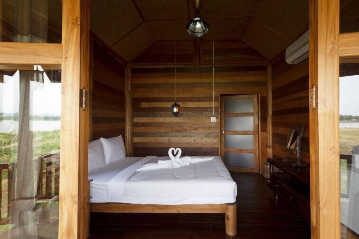 A wooden cabin room with a bed, a small table, and a window overlooking a natural landscape.