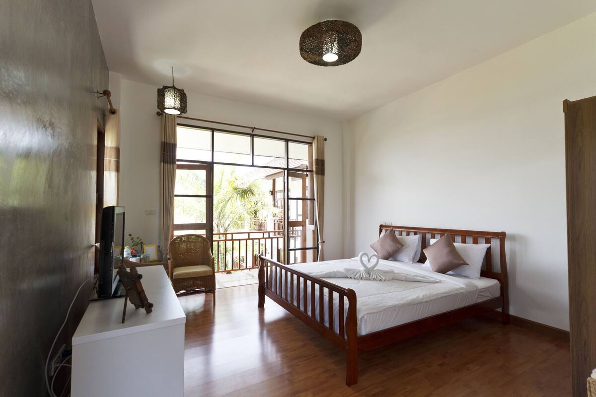 A bedroom with a wooden bed, a white bedside table, a television, and a large window with a view of greenery.