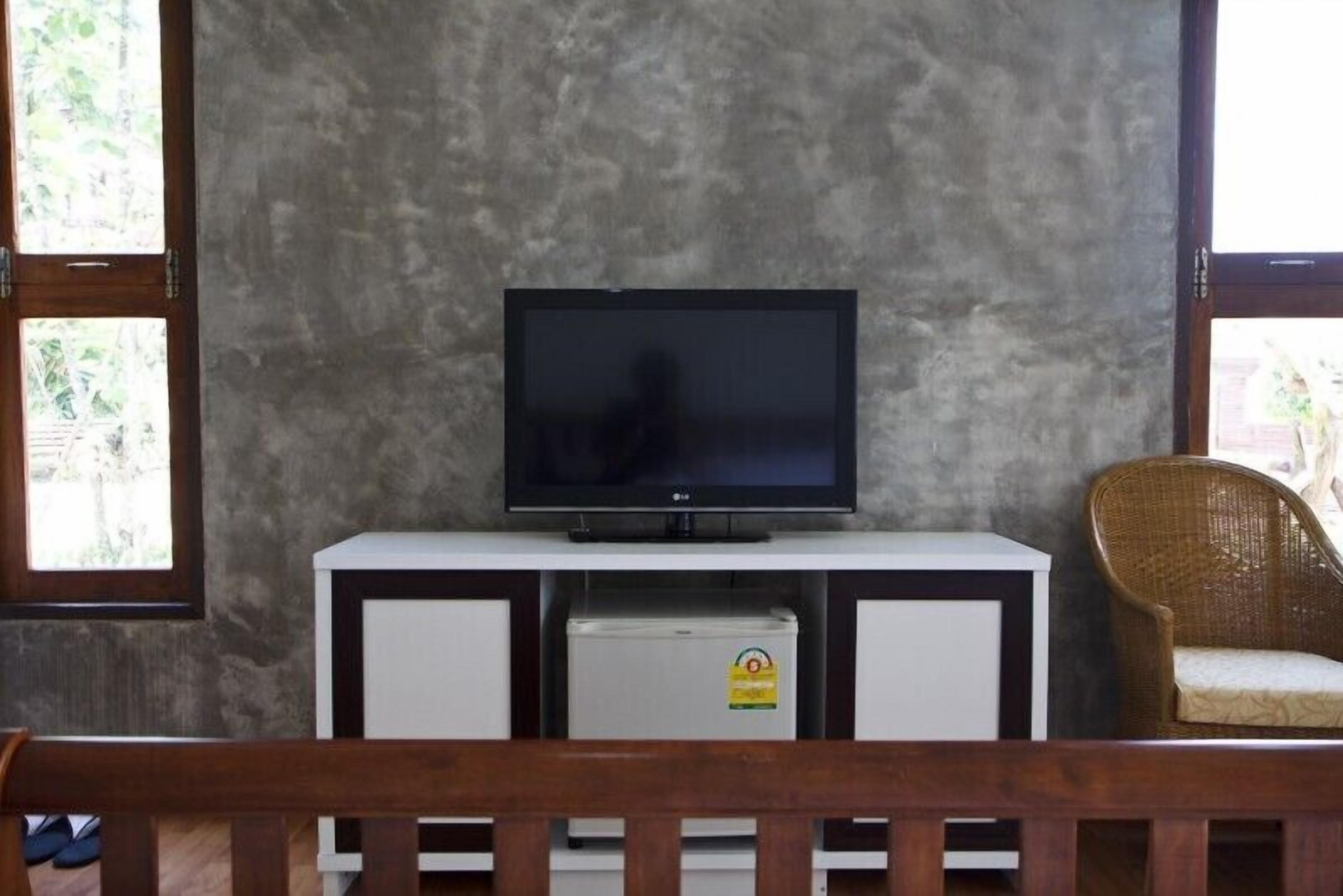 A television on a white cabinet with a small refrigerator below it, a wicker chair to the right, and a wooden bench in the foreground.