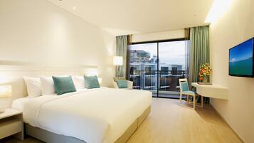 Ein Hotelzimmer mit einem großen Bett, einem Schreibtisch mit Stuhl, einem Fernseher und einem Balkon mit Blick auf Gebäude.