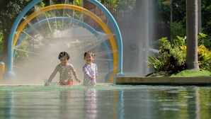 Two children playing in a water park with a large water fountain and greenery in the background.