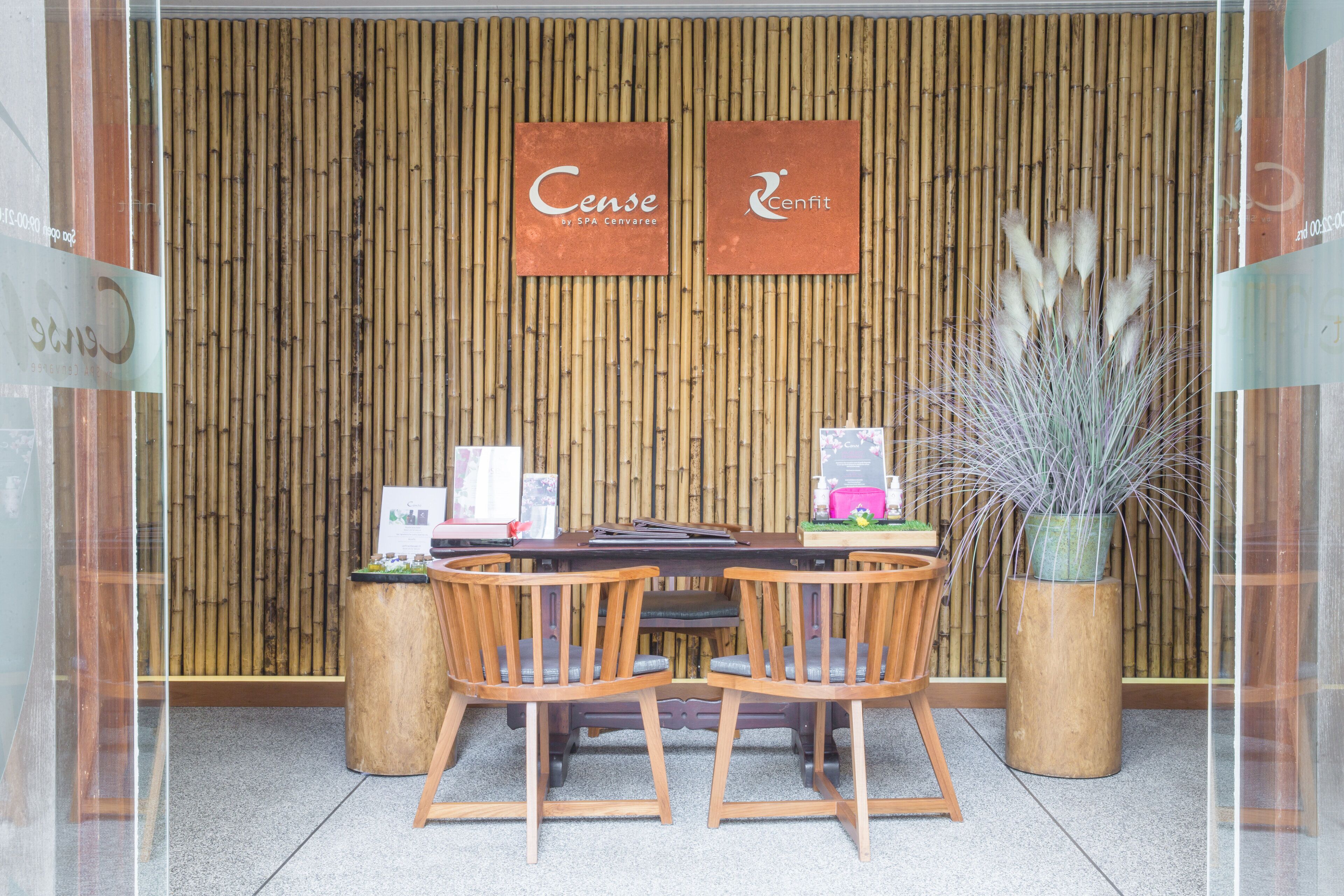 Un área de recepción con una pared de bambú, dos letreros que dicen 'Cense' y 'Cenfit', un mostrador de recepción, dos sillas de madera y una planta en maceta.