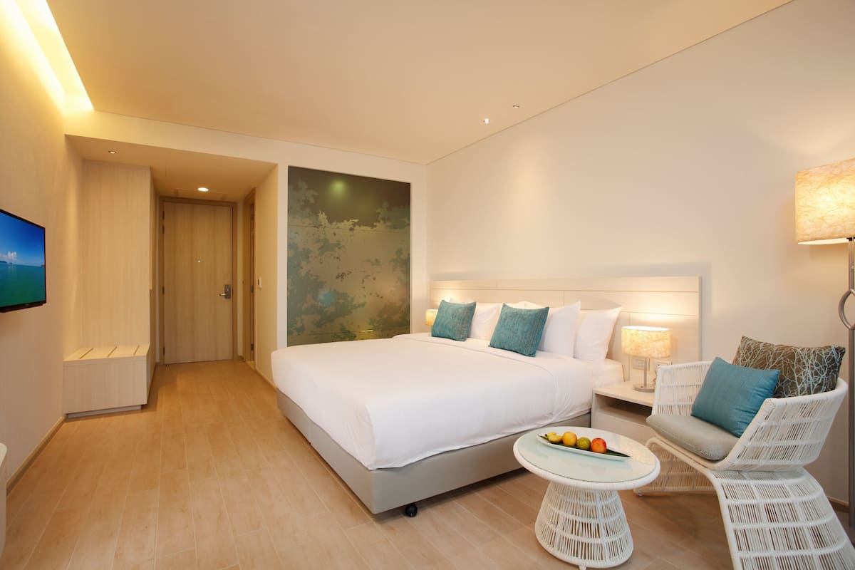 A modern hotel room with a large bed, a small round table with fruit, a white armchair, and a TV on the wall.