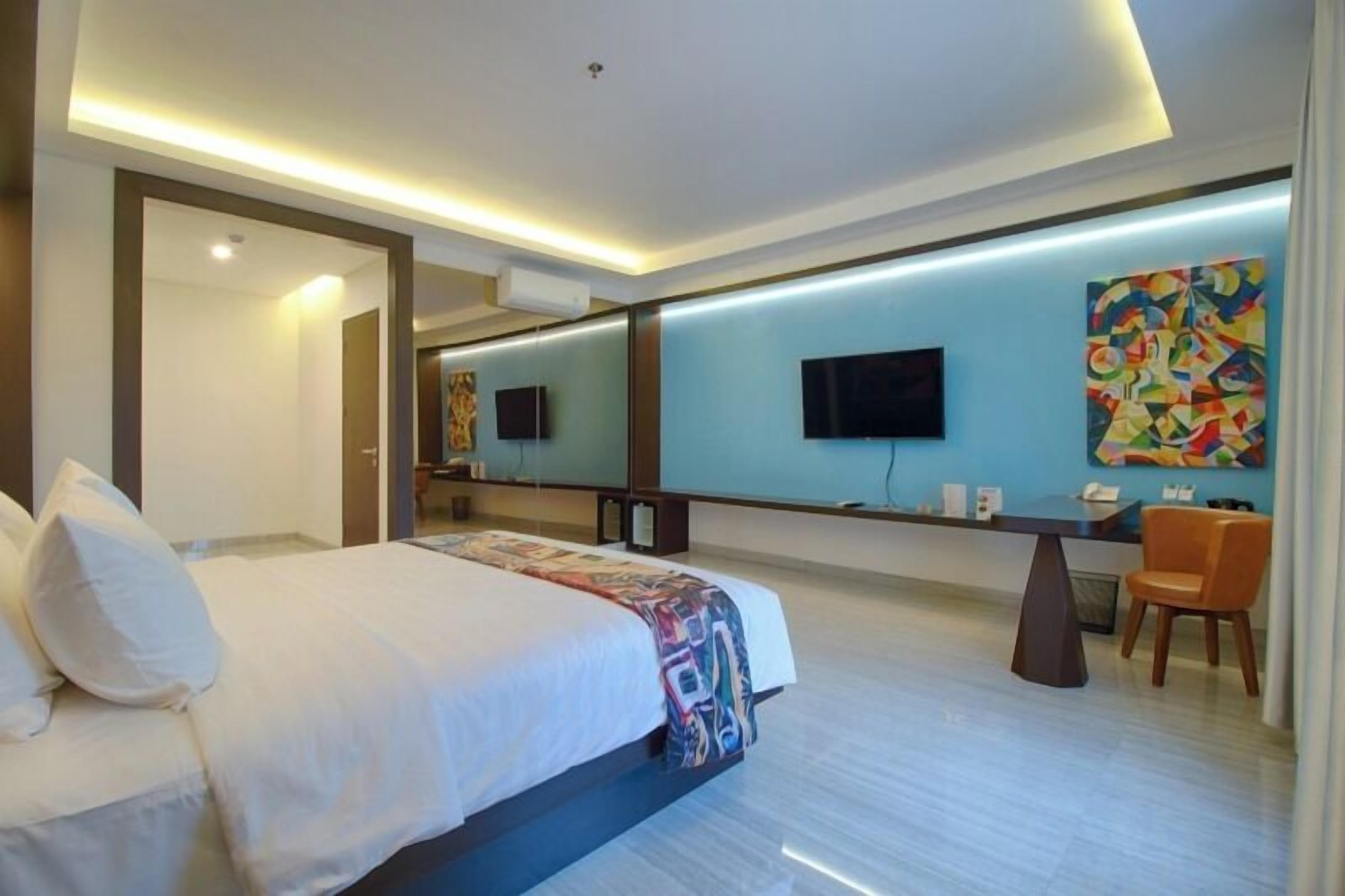 A modern hotel room with a large bed, a desk, a chair, a flat-screen TV, and a colorful abstract painting on the wall.