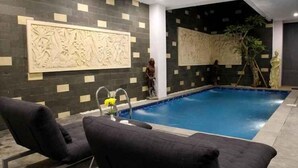 Un área de piscina interior moderna con paredes de azulejos oscuros, una piscina central y dos sillas de descanso negras.