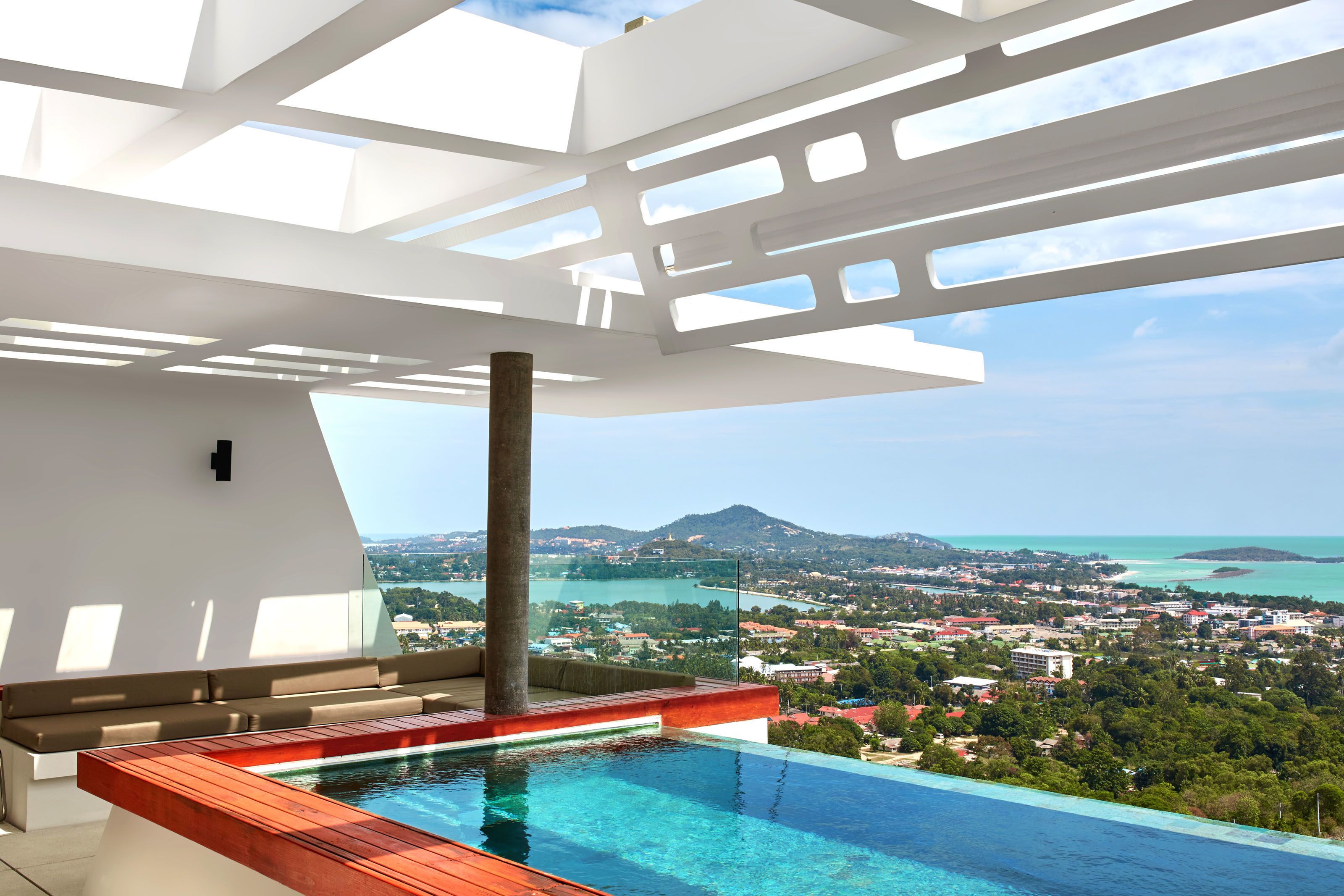 A modern infinity pool with a wooden deck, a white ceiling with skylights, and a view of a coastal town and mountains.