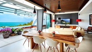 A modern living room with a large wooden dining table, white chairs, a sofa, and a TV. There are potted plants and a view of the ocean. - Aqua Samui Villas (Koh Samui)
