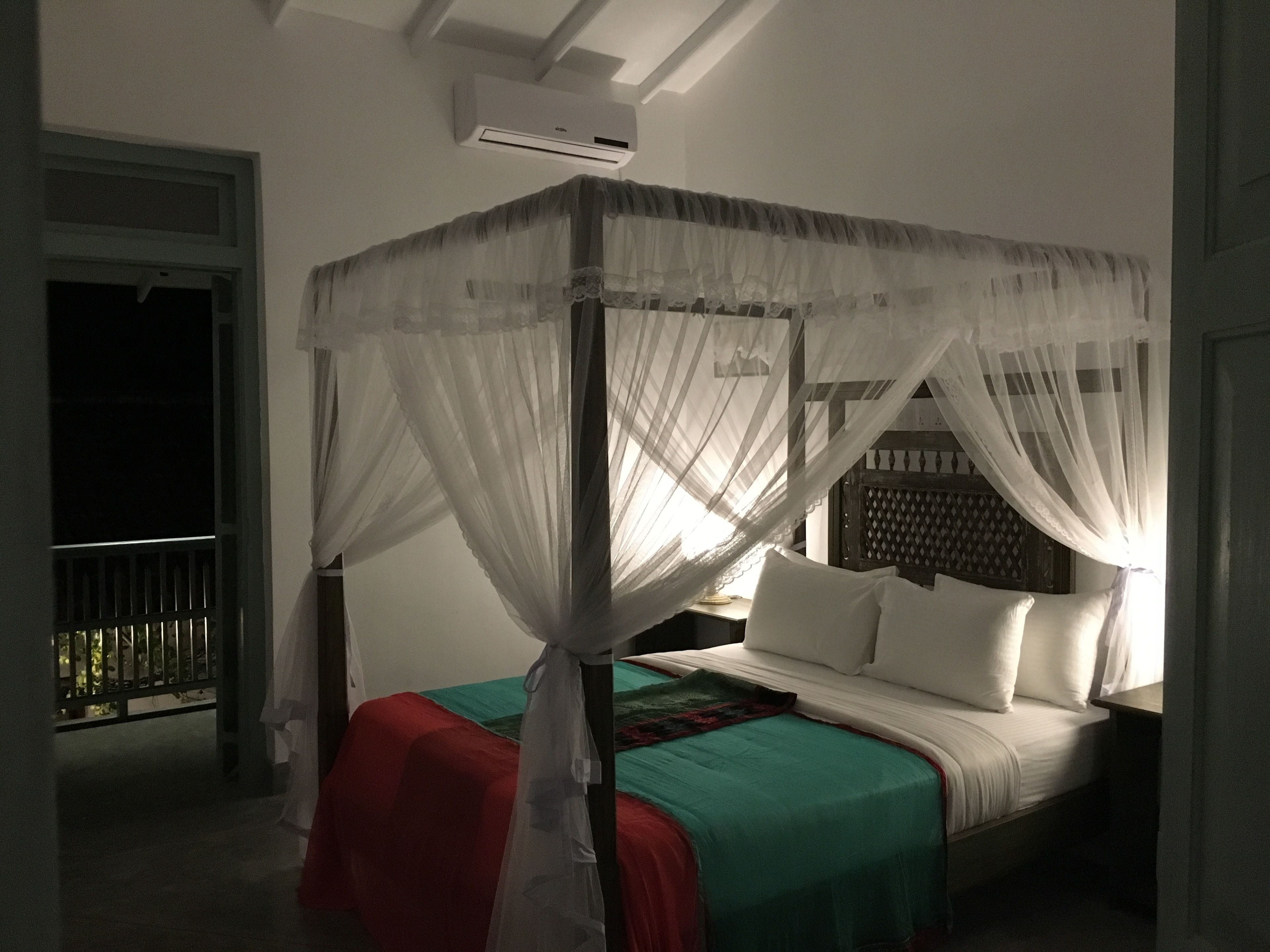 A four-poster bed with white bedding and a red blanket, an air conditioning unit above, and a balcony with a metal railing.