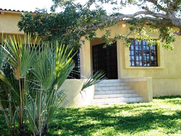 Un edificio amarillo con puerta y ventana negras, rodeado de césped verde y una palmera.