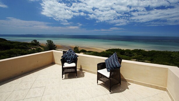 A balcony with two wicker chairs, each with a white cushion and a blue pillow, overlooking a beach with clear turquoise waters and a sandy shore under a partly cloudy sky. - Deacra Villas By Sol Resorts (Vilanculos)