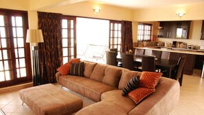 A living room with a large sectional sofa, a dining area with a table and chairs, and a kitchen in the background. - Deacra Villas By Sol Resorts (Vilanculos)