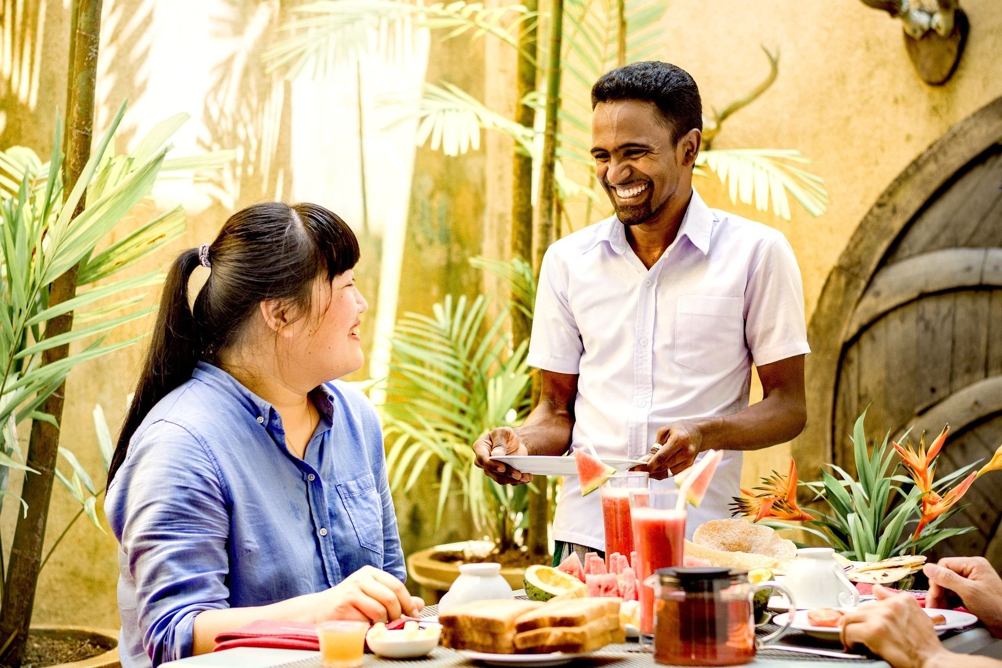A dining area with a table set for a meal, surrounded by tropical plants and a rustic wall.