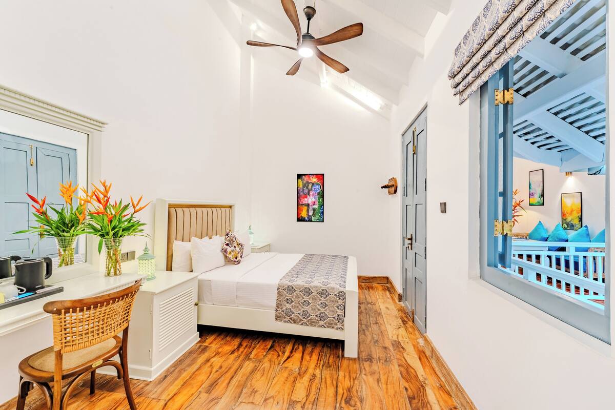 Une chambre à coucher comprenant un lit, un bureau, une chaise, un ventilateur de plafond et donnant sur un balcon extérieur.