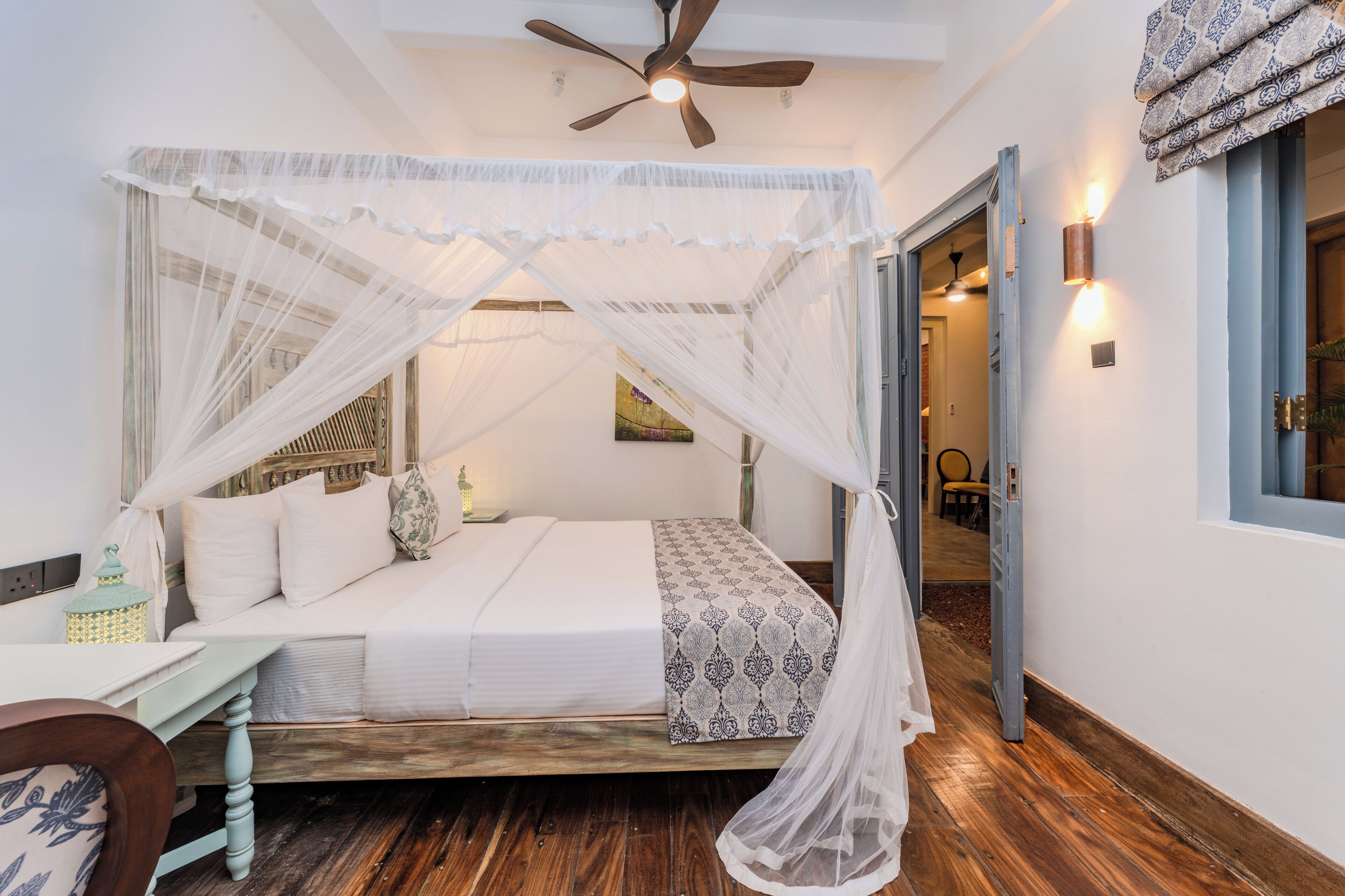 A room with a bed under a canopy, a wooden floor, a small table with a lamp, and a chair.