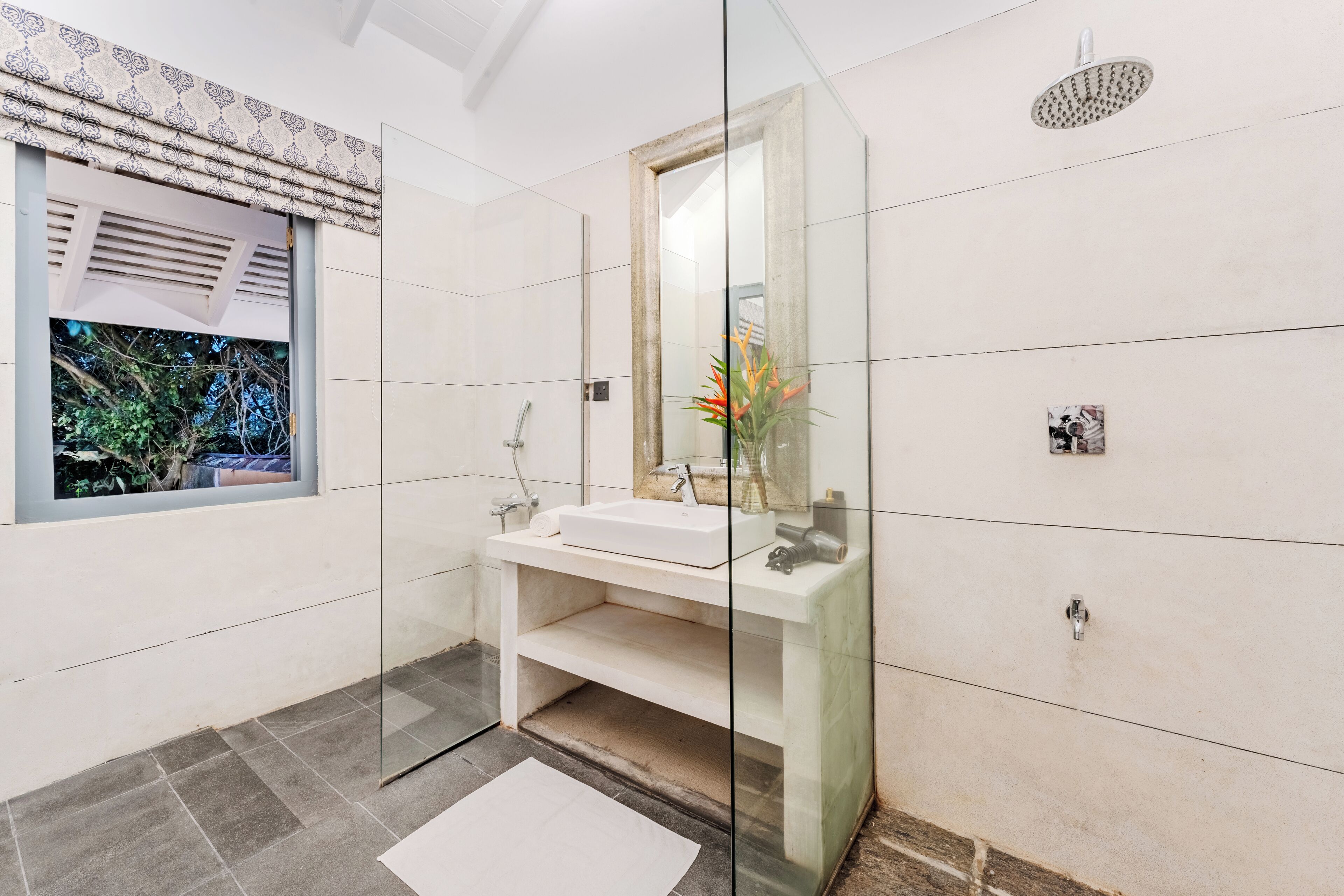 A modern bathroom with a glass shower enclosure, a white sink, and a window with a view of greenery.