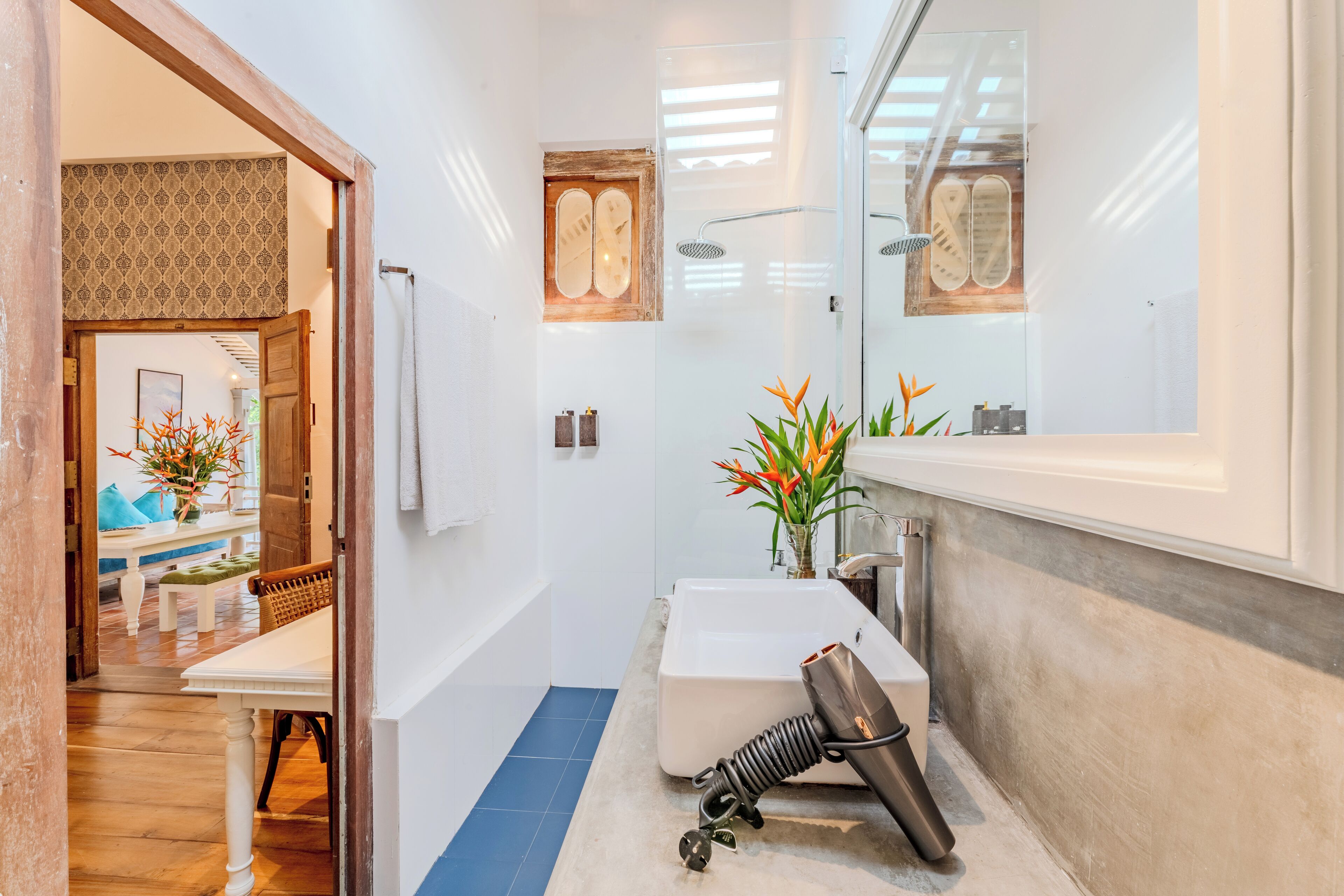 A modern bathroom with a large mirror, a white sink, and a shower. There is a towel rack and a bench in the adjacent room.
