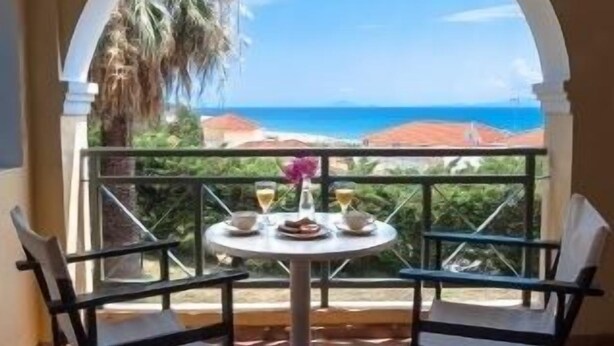 Un balcón con una mesa dispuesta para dos, con vistas a la costa, palmeras y cielo despejado.