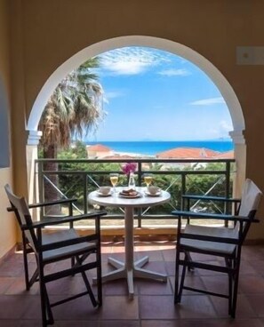 A balcony with a table set for two, overlooking a coastal view with palm trees and a clear sky.