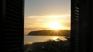 Vista desde una ventana con contraventanas, con atardecer sobre una masa de agua y una forma de terreno distante.