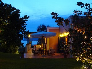 An outdoor patio area with a covered seating arrangement, a view of the sea, and a building with warm lighting. - Mounda Beach Hotel (Kefalonia)