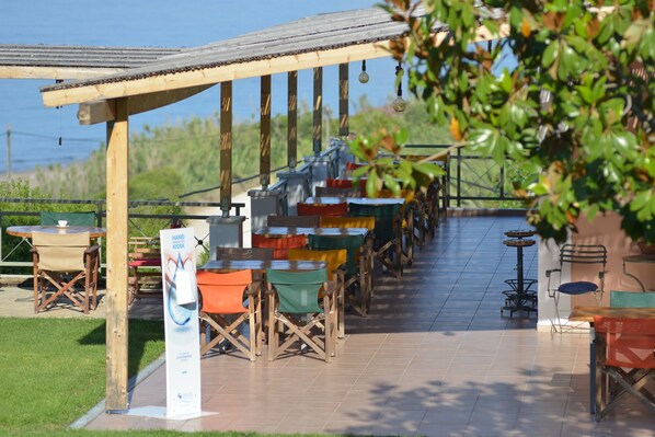 An outdoor seating area with multiple tables and chairs, a covered section, and a kiosk. - Mounda Beach Hotel (Kefalonia)