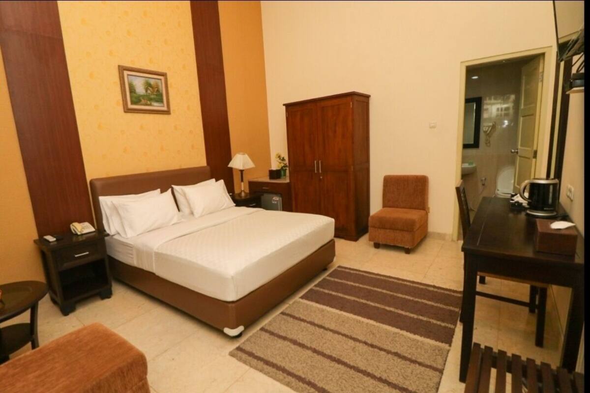A hotel room with a bed, a wooden wardrobe, a desk with a chair, a small table, and a nightstand.
