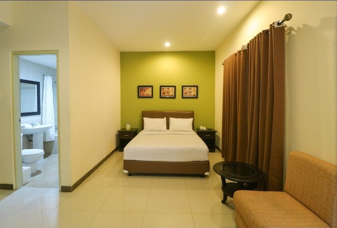Habitación de hotel con cama, mesita de noche, una pequeña mesa, un sofá y un baño visible a través de una puerta abierta.