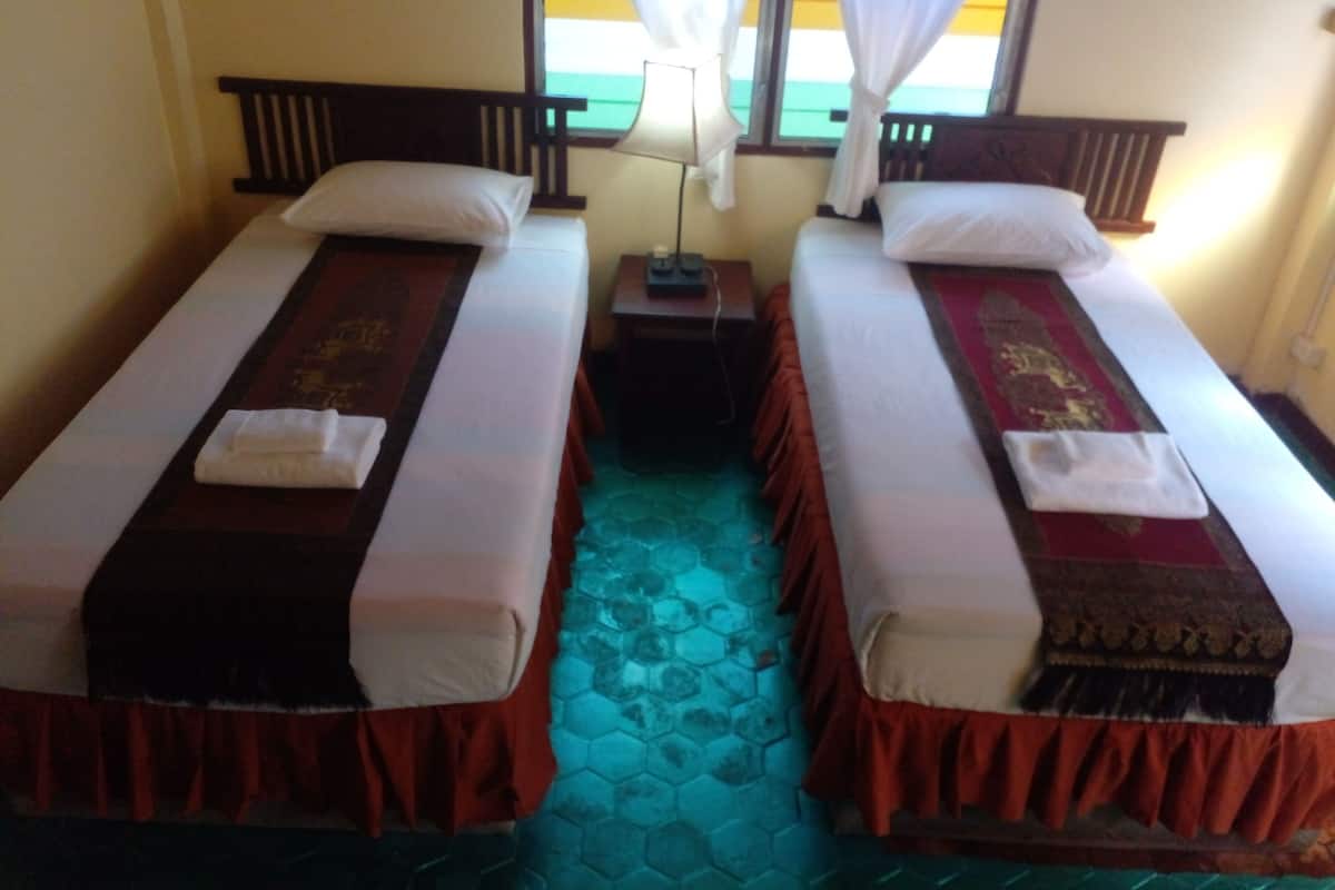 Two single beds with red and brown bedspreads, a small bedside table with a lamp, and a window with white curtains.