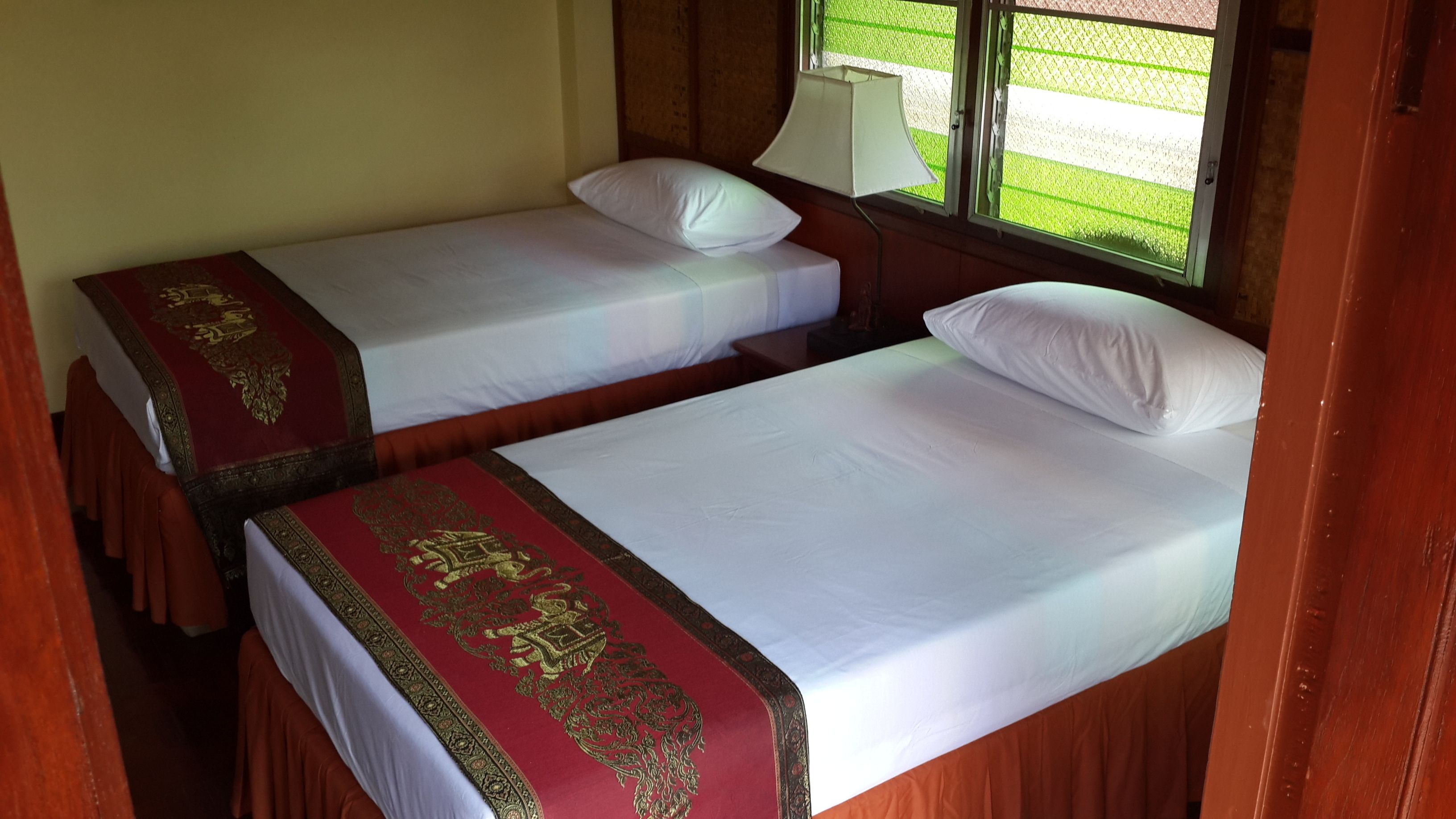 Two single beds with red and gold bedspreads, a window with a view of a green field, and a lamp on the nightstand.
