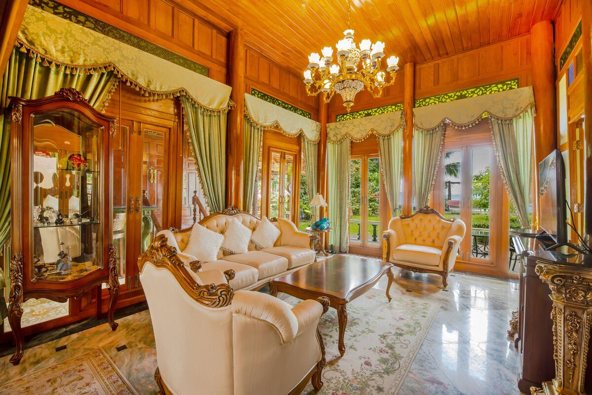 Une pièce spacieuse avec un grand canapé, un fauteuil plus petit, une table basse et un téléviseur. Un lustre est suspendu au plafond et un grand miroir orne le mur.
