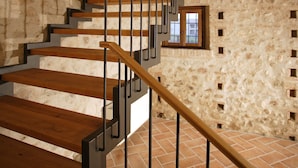 Una escalera de madera con barandales metálicos que conduce a un nivel superior con un muro de piedra y una ventana pequeña.