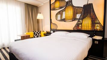 Quarto de hotel com uma cama grande, uma mesa com luminária e uma obra de arte na parede.