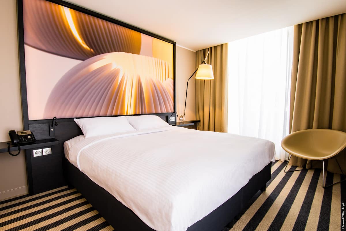 A hotel room with a large bed, a chair, a nightstand with a phone, and a wall-mounted lamp.