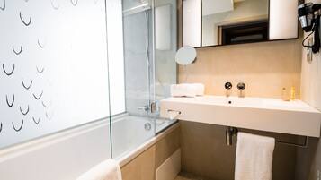 A modern bathroom with a glass shower enclosure, a white sink with a mirror above, and two towels on the floor.