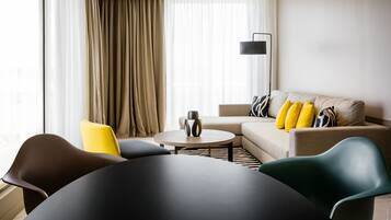 Een moderne woonkamer met een ronde tafel, een bank met gele kussens en een groot raam met dunne gordijnen.