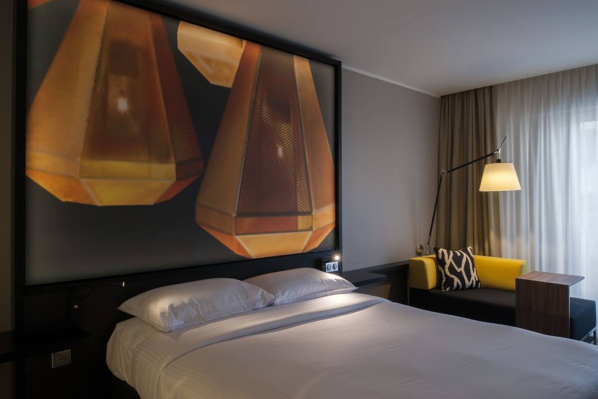 A hotel room with a large bed, a nightstand, a lamp, and a sofa with a patterned pillow.