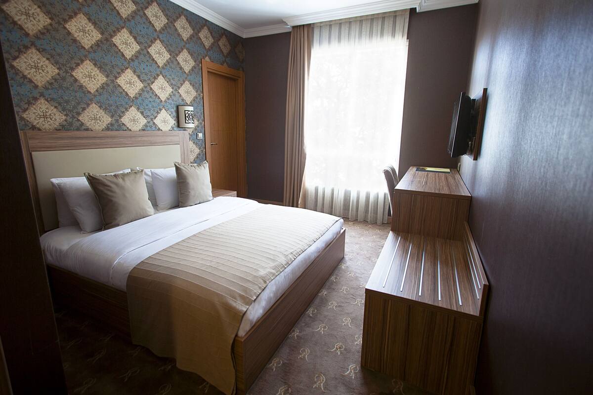 A hotel room with a large bed, a wooden nightstand, a TV mounted on the wall, and a patterned wallpaper.