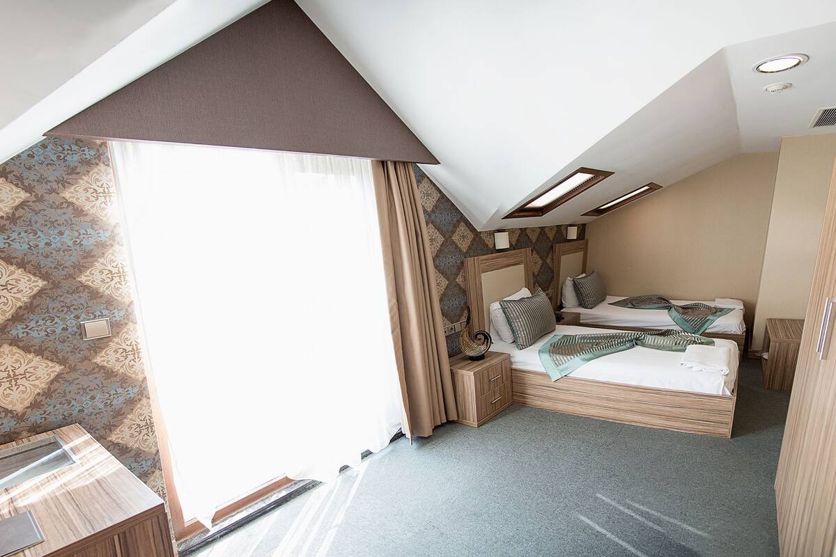 A bedroom with a sloped ceiling, patterned wallpaper, a bed with pillows, a bedside table, and a small wooden cabinet.